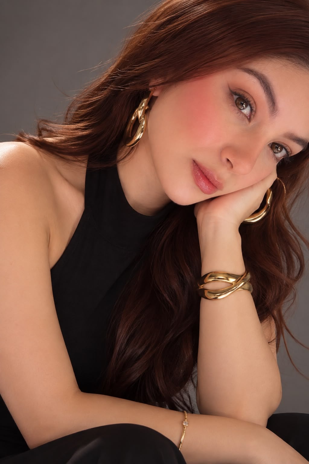 Angelica Barreto, maquilladora profesional, con el cabello largo castaño, blush rosado, pendientes y brazaletes dorados y la mejilla apoyada en la mano