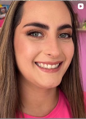Mujer sonriente de ojos verdes con cejas definidas, rubor rosado y labios en tono natural, con el cabello largo castaño y una blusa rosada