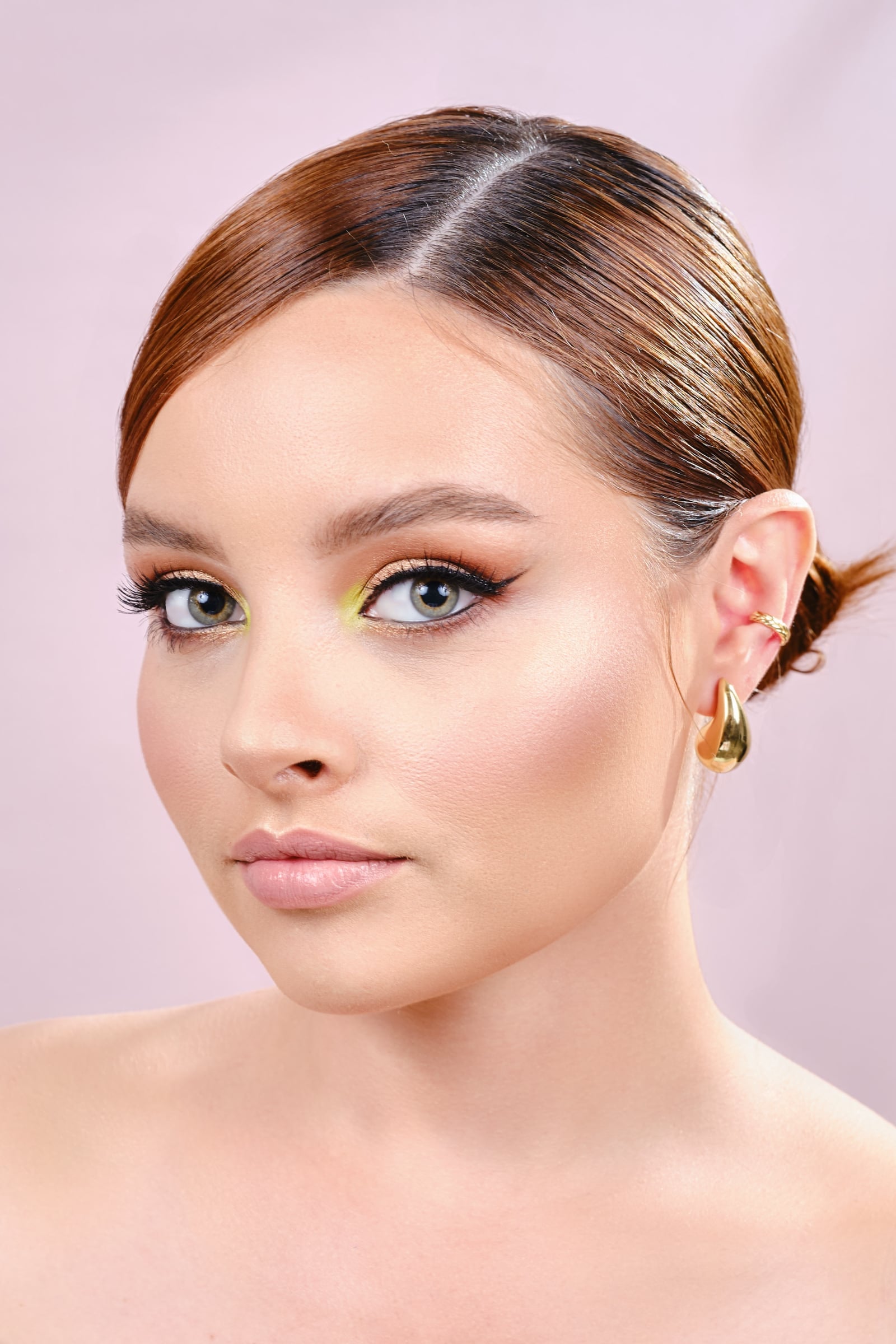 Peinado pulido con el cabello recogido hacia atrás y un moño bajo, con maquillaje de sombra verde y amarilla y delineado alado, sobre fondo rosa