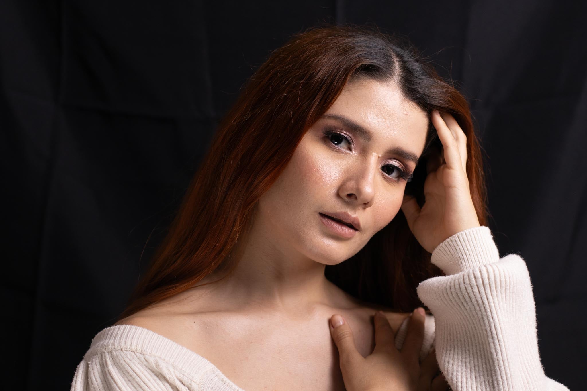 Maquillaje natural de estudio: mujer de cabello cobrizo con una mano en el cabello, piel luminosa, rubor cálido y labios rosados, sobre fondo negro