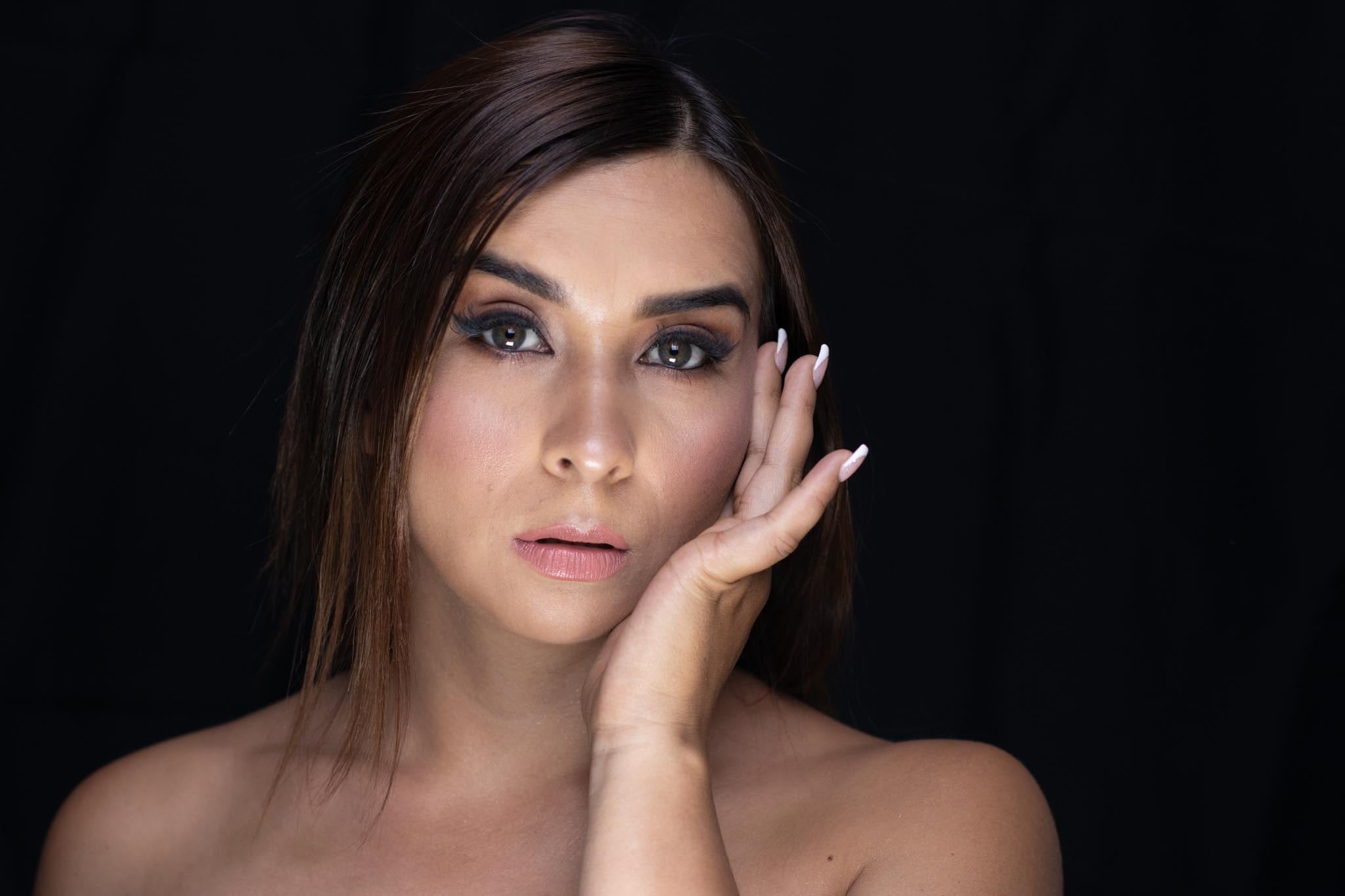 Retrato con la mano junto a la mejilla, mirada al frente, ojos definidos y labios nude, sobre fondo negro