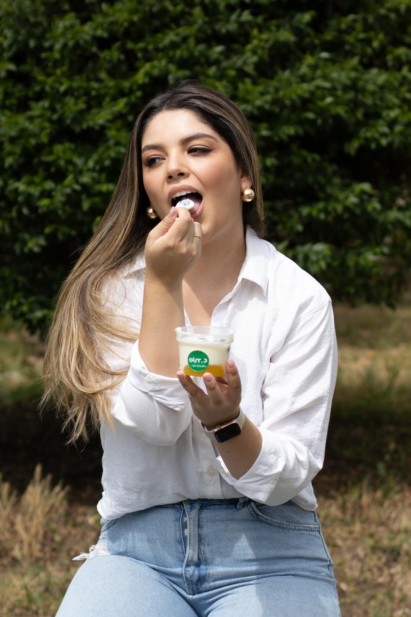 Maquillaje fresco y natural al aire libre: mujer con camisa blanca y jeans, cabello largo con reflejos, que prueba un postre con una cuchara frente a un seto verde