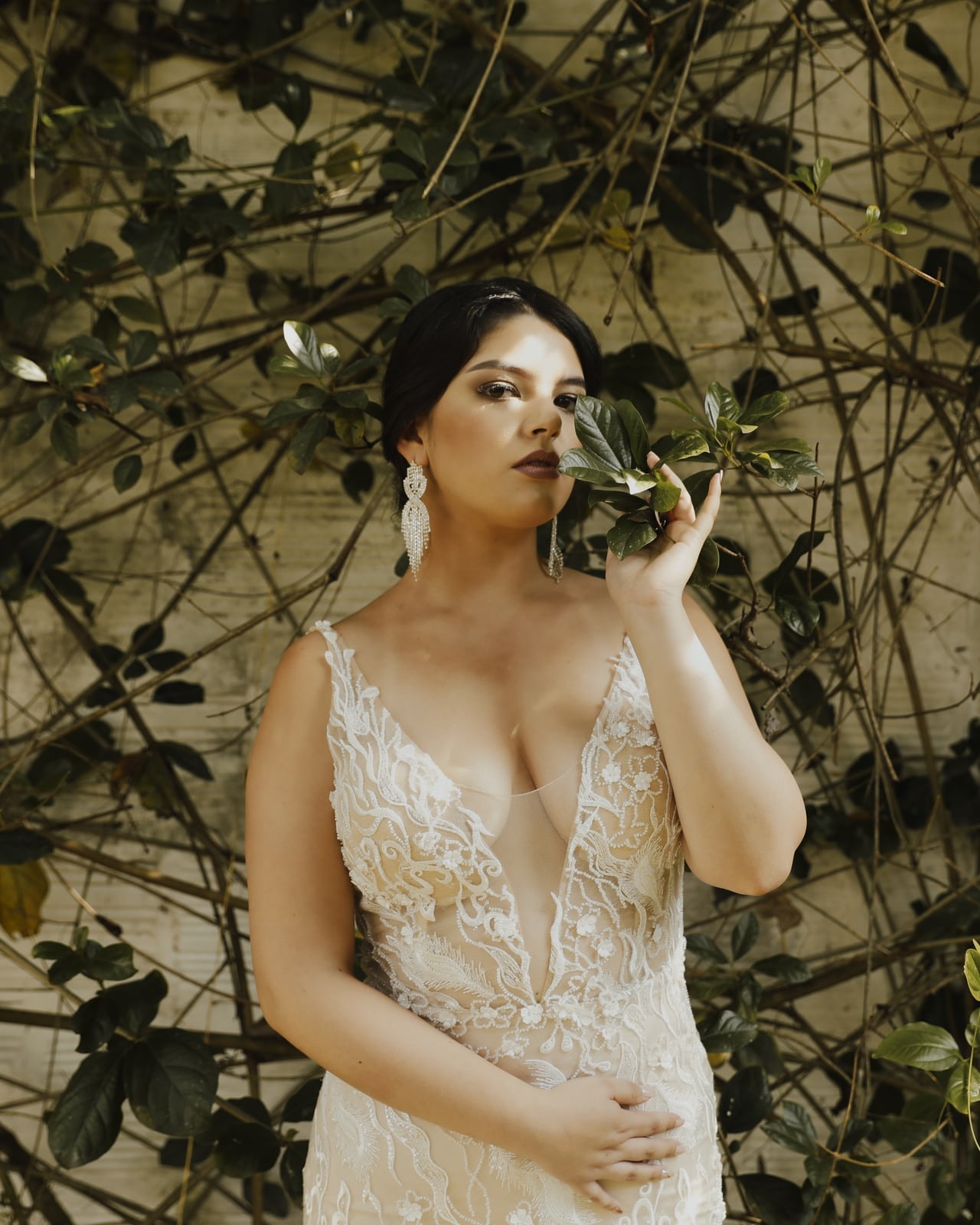 Novia con vestido de encaje y aretes largos, con una hoja junto al rostro, frente a un muro con enredadera